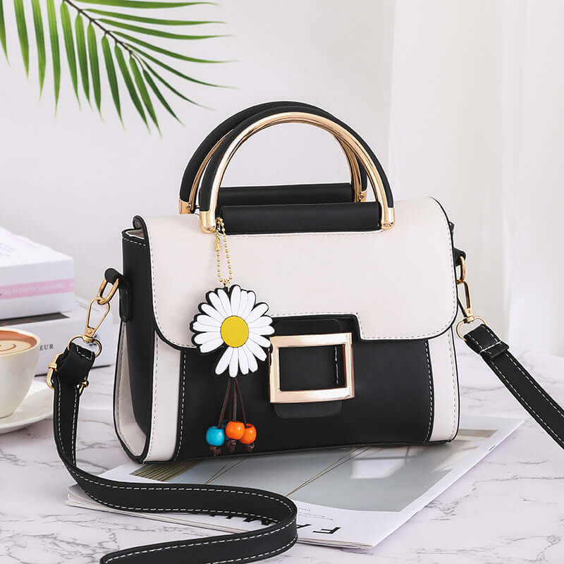 Stylish women's crossbody bag in black and white with floral charm and gold accents on a desk.
